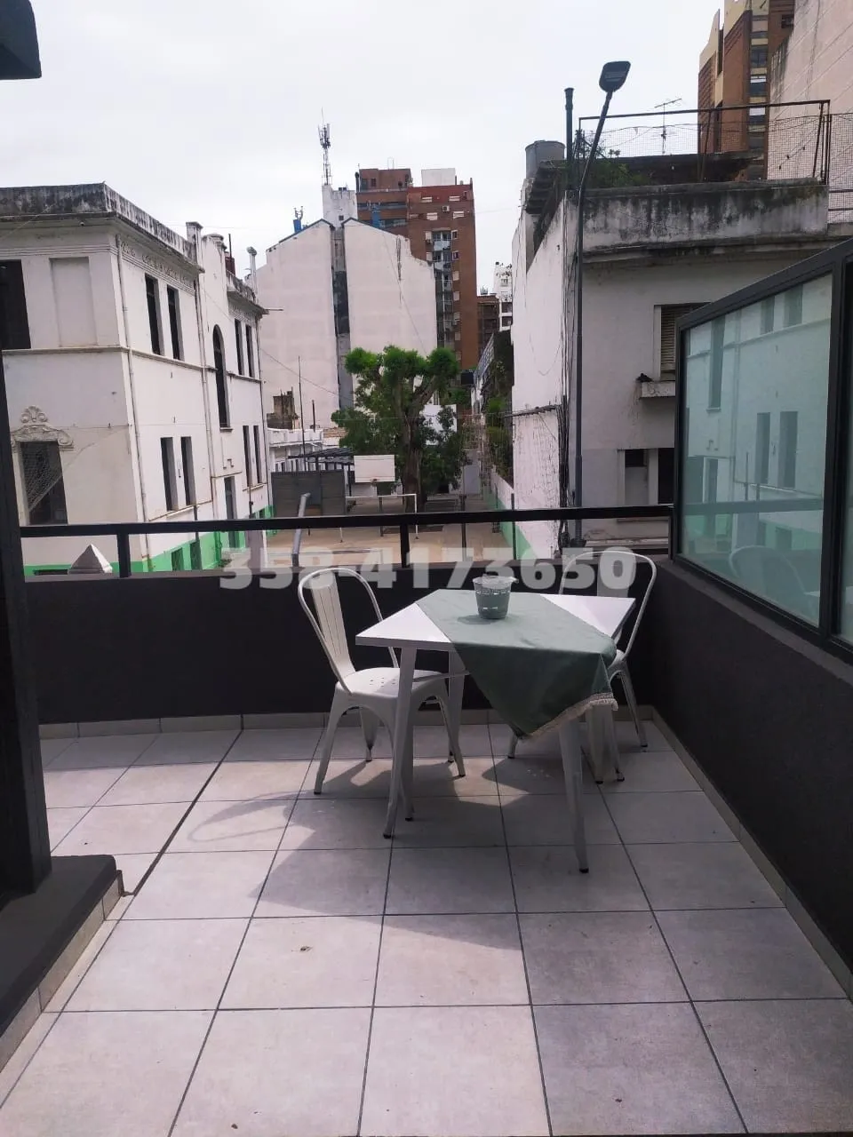 Balcon con mesa blanca cubierta con mantel verde y dos sillas metálicas blancas, edificios alrededor y árbol al fondo