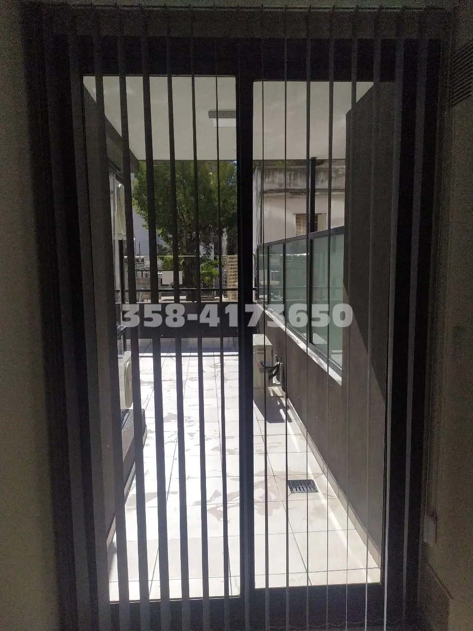 Balcón pasillo con barandas metálicas verticales y paneles de vidrio, vista hacia el exterior con árboles y edificios vecinos.