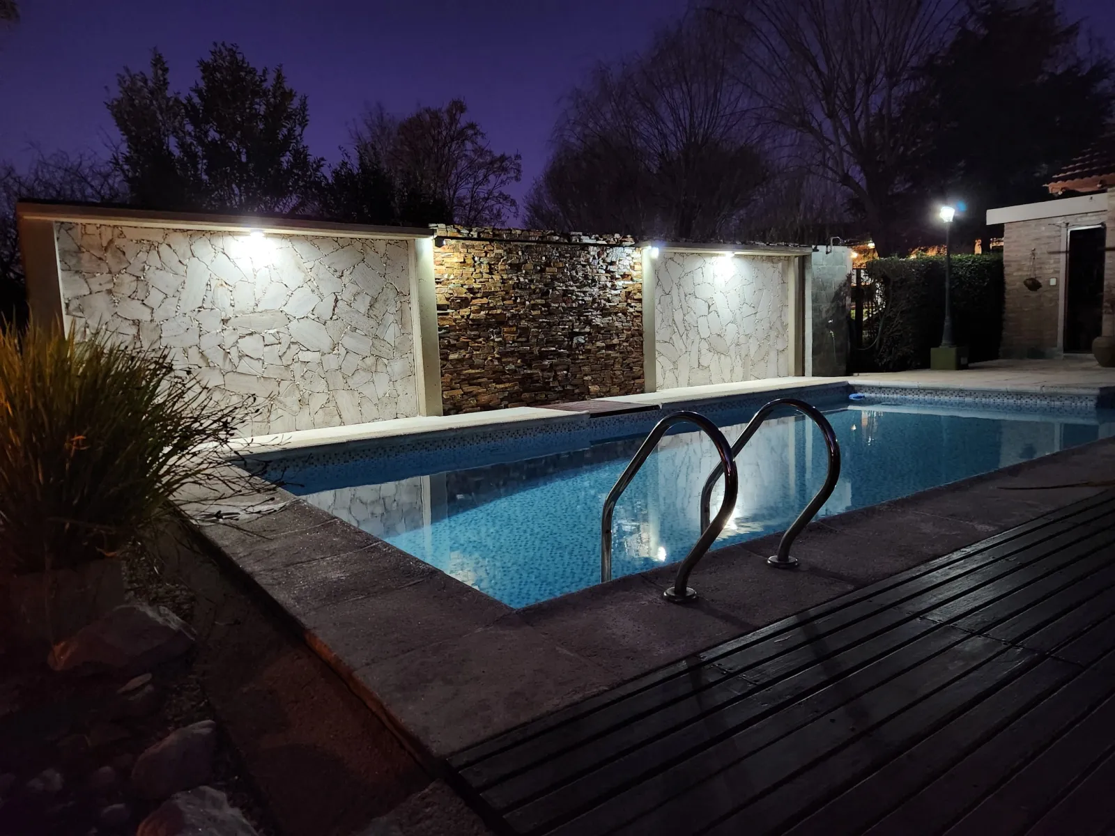 Pileta rectangular con escalera metálica, luz en paredes de piedra y área con deck de madera al anochecer