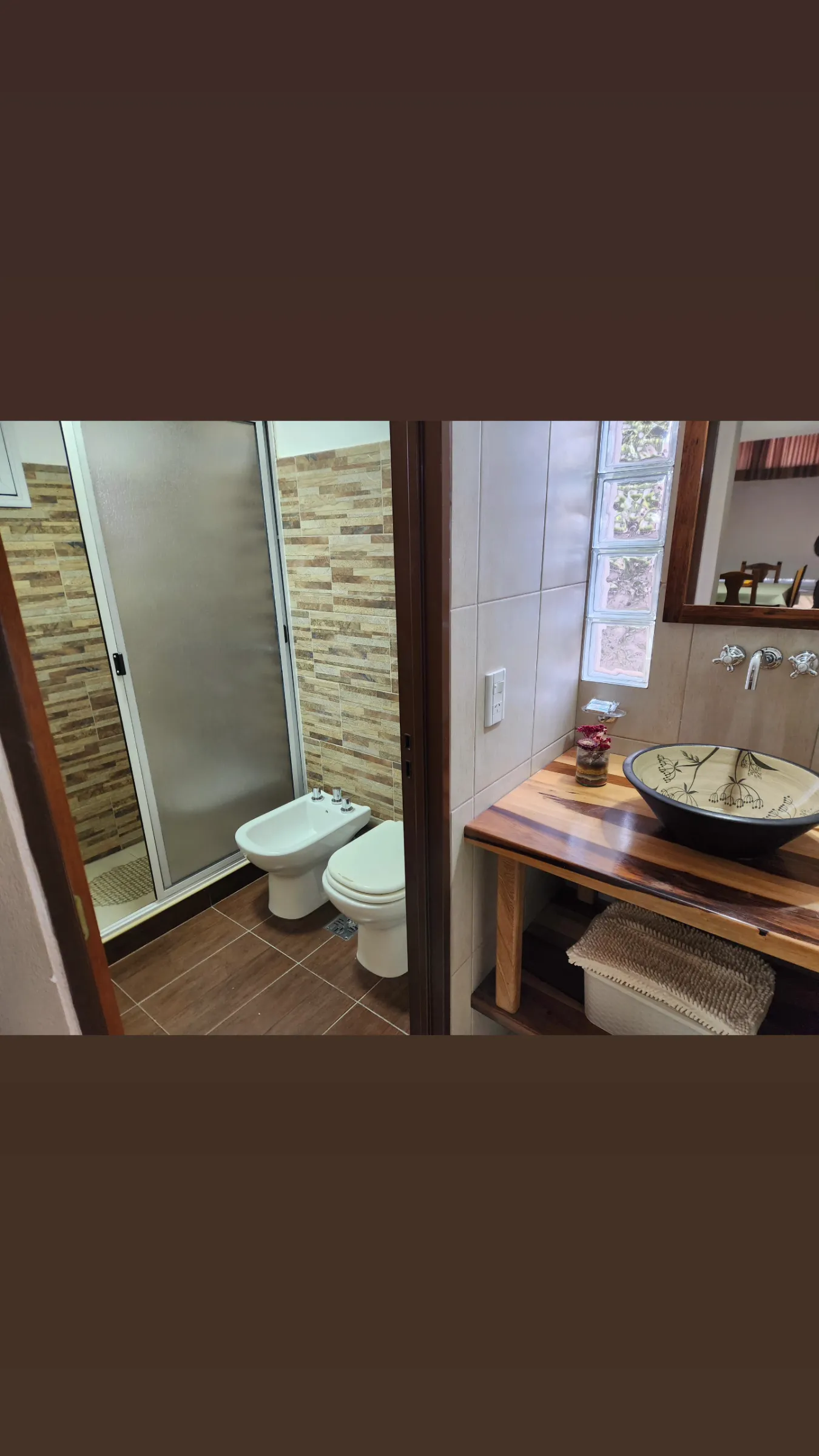 Interior de un baño con ducha con mampara, bidet, inodoro, y bacha de cerámica sobre mueble de madera con espejo en la pared