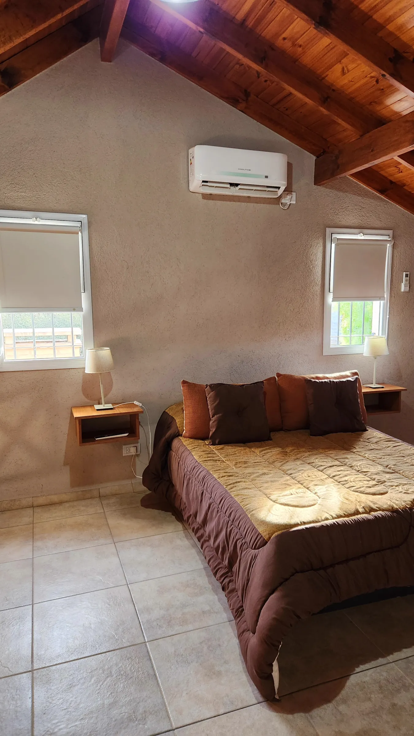 Habitación con una cama doble cubierta con acolchado marrón claro y varias almohadas marrones, dos ventanas con cortinas enrollables y dos mesas de apoyo con lámparas.