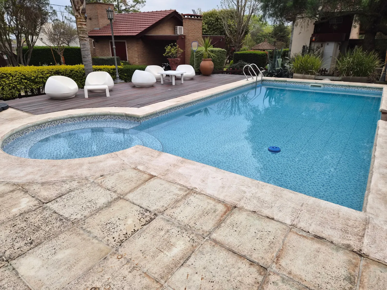 Pileta de natación con borde de piedra y espacio de madera con sillones blancos y mesa redonda