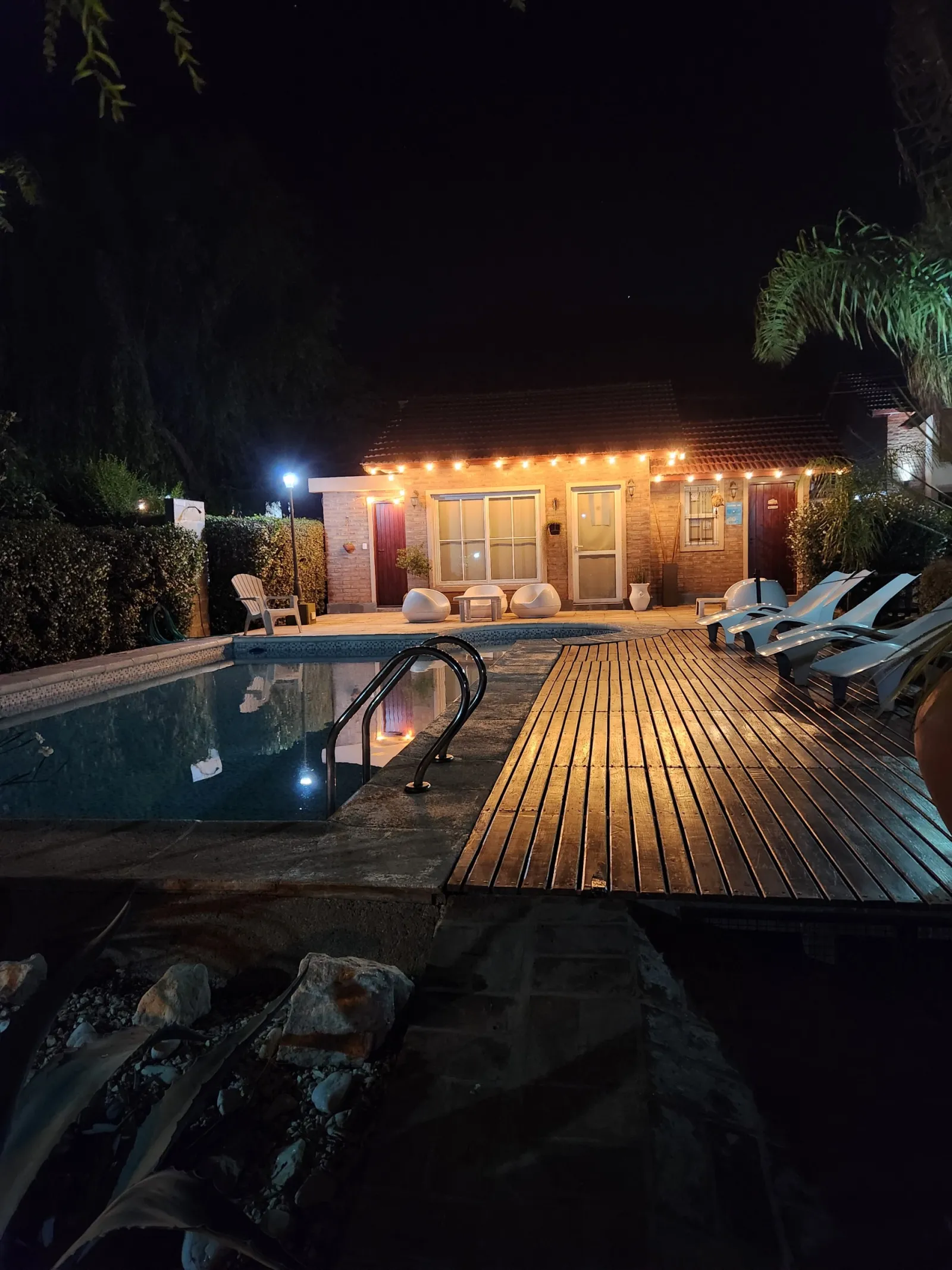 Piscina con escalera, terraza de madera con reposeras y sillones frente a una casa iluminada con luces cálidas