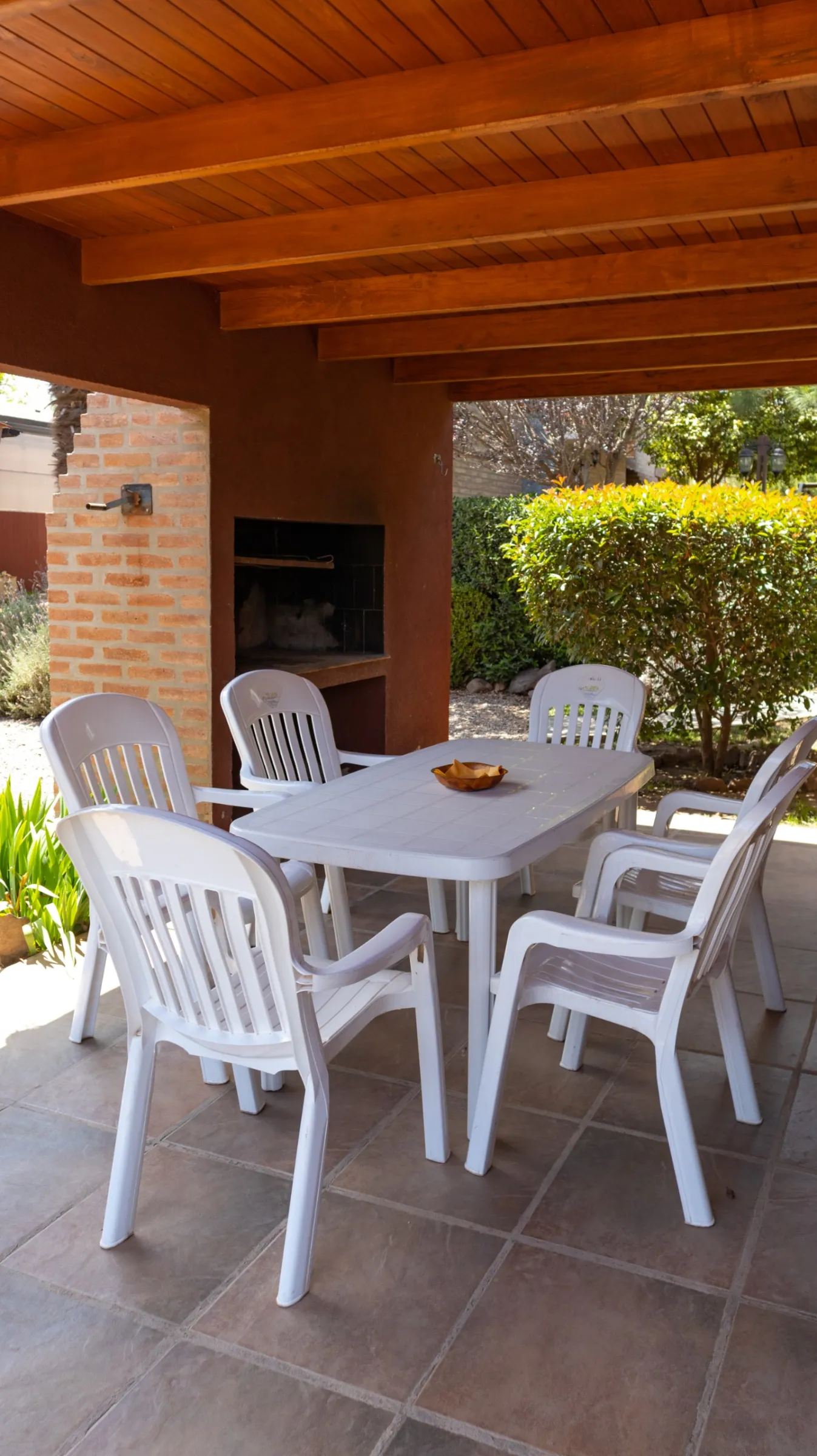 Patio con mesa plástica rectangular blanca con seis sillas blancas alrededor y asador de ladrillo a la vista